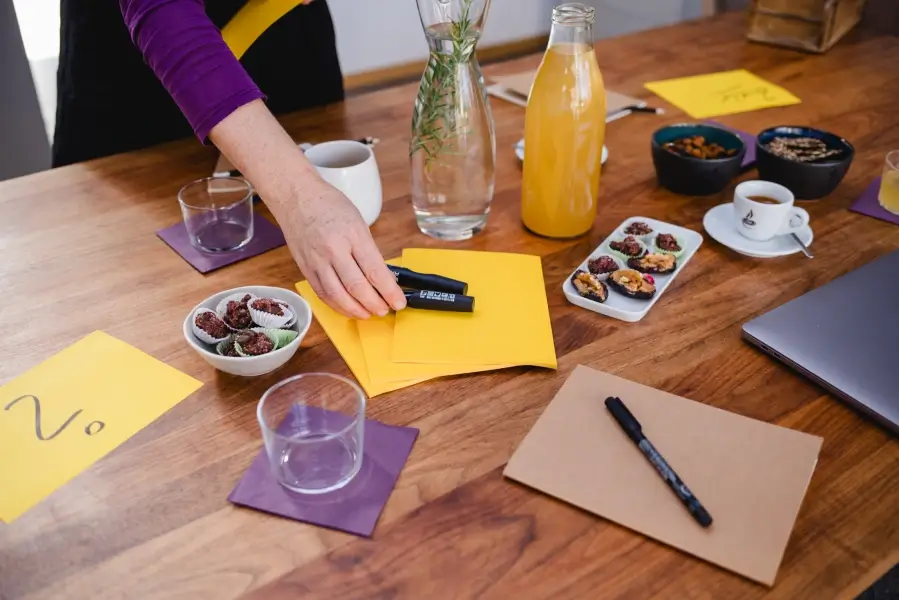 Workshop in der Concept Kitchen wird vorbereitet. Es liegen Zettel und Stifte auf dem Tisch. Eine Hand platziert weitere Stifte. Als Verpflegung stehen Rosmarinwasser, Limonade, Brainfood und Espresso auf dem Tisch